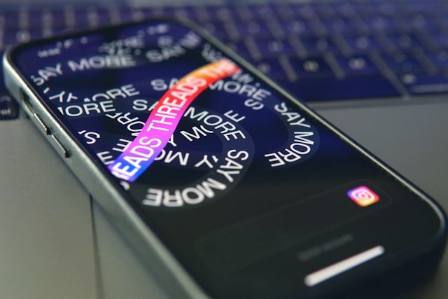 A smartphone displaying the Threads app screen with colorful text that says "SAY MORE" and "THREADS."