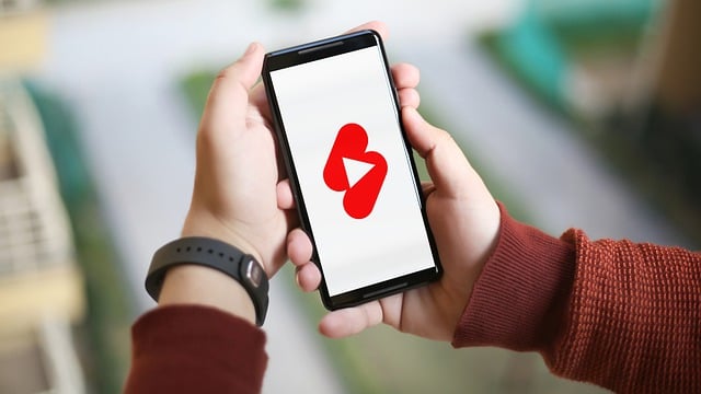 A person holding a smartphone displaying the YouTube Shorts logo on the screen, with a blurred background.