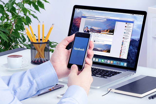 A person holding a smartphone with the Facebook app open, with a laptop showing a Facebook profile on a desk.