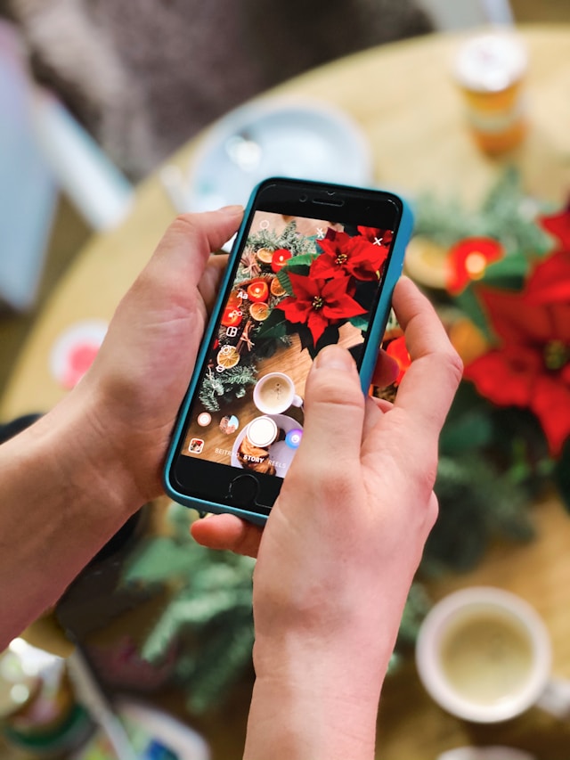 A person capturing a festive image of red poinsettias, dried oranges, and a cup of coffee on a table for their Instagram story.