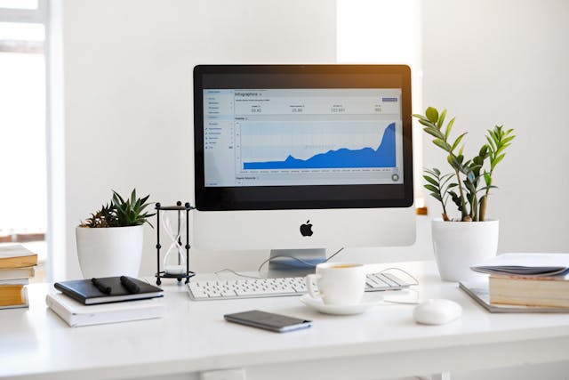 A clean desk setup with a computer displaying a growth analytics chart, surrounded by plants, notebooks, and a coffee cup.