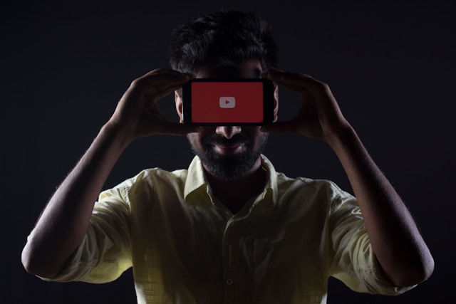 A person holding a smartphone with YouTube play button covering their eyes, against a dark background.