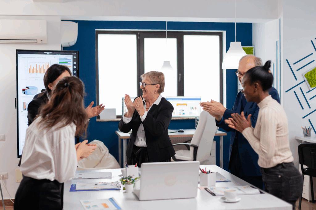 A group of people stand around the table in the office and applaud each other, looking at the chart on the big screen.