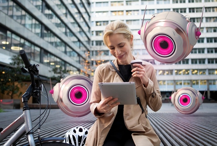 woman with a tablet and coffee sits near her bike while futuristic floating robots hover around her.
